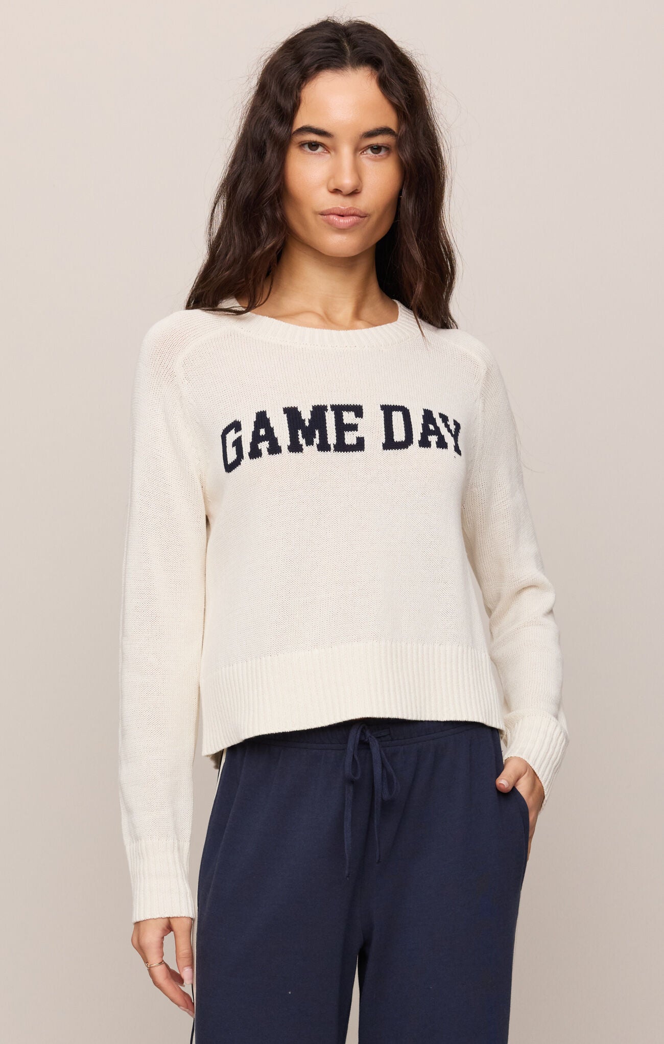 Woman wearing a white sweater with 'GAME DAY' text and navy pants on a plain background