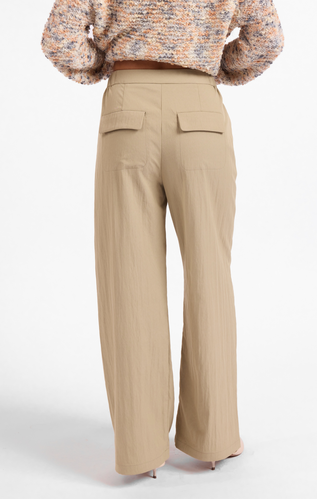 A person shown from the back wearing beige wide-leg trousers with two flap back pockets, paired with a textured multicolor knit top and heels, against a plain light gray studio background.
