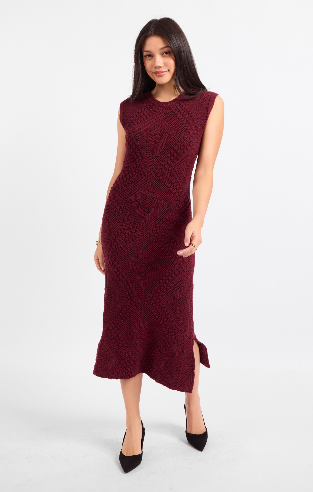 A woman modeling a sleeveless, crew-neck midi dress in deep burgundy with a textured, raised knit pattern and a side slit, paired with black pointed-toe heels against a light gray studio background.