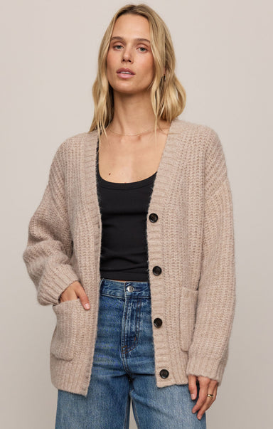 Woman wearing a beige cardigan over a black top and blue jeans on a plain background