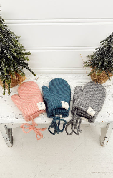 Three pairs of colorful mittens on a white bench with Christmas trees in the background.