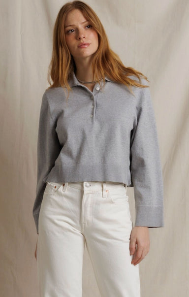 A woman modeling a light gray long-sleeve knit polo shirt with white high-waisted jeans, standing against a neutral beige fabric backdrop.