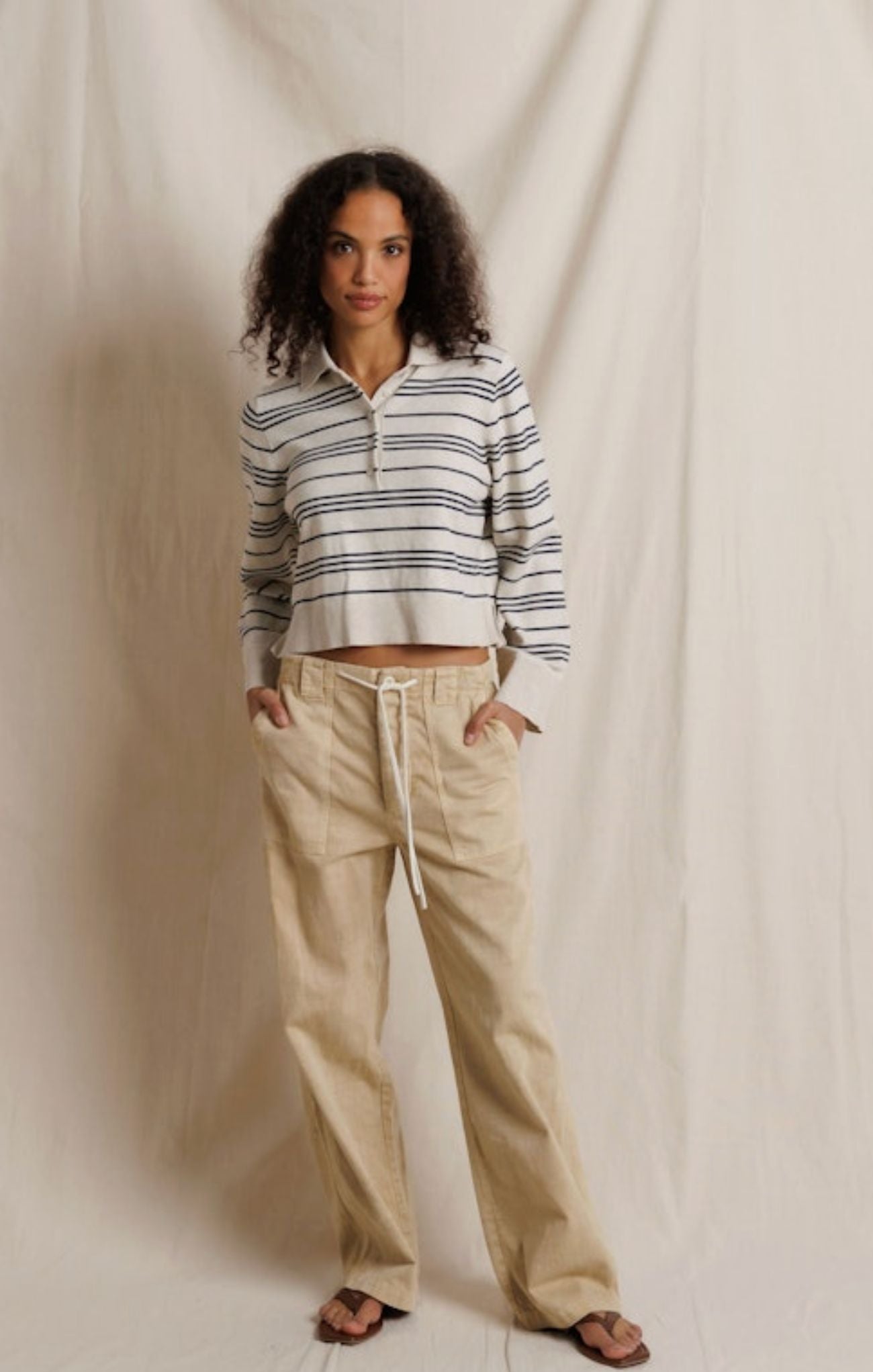 A woman stands against a draped cream fabric backdrop wearing a cropped long-sleeve polo-style top with thin dark horizontal stripes and beige wide-leg drawstring pants.