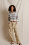 A woman stands against a draped cream fabric backdrop wearing a cropped long-sleeve polo-style top with thin dark horizontal stripes and beige wide-leg drawstring pants.