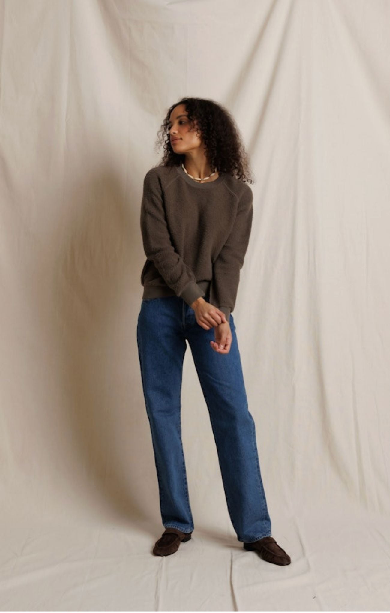 A woman modeling a dark brown crewneck sweater and blue straight-leg jeans against a draped off-white fabric backdrop.