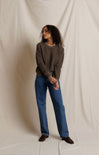 A woman modeling a dark brown crewneck sweater and blue straight-leg jeans against a draped off-white fabric backdrop.