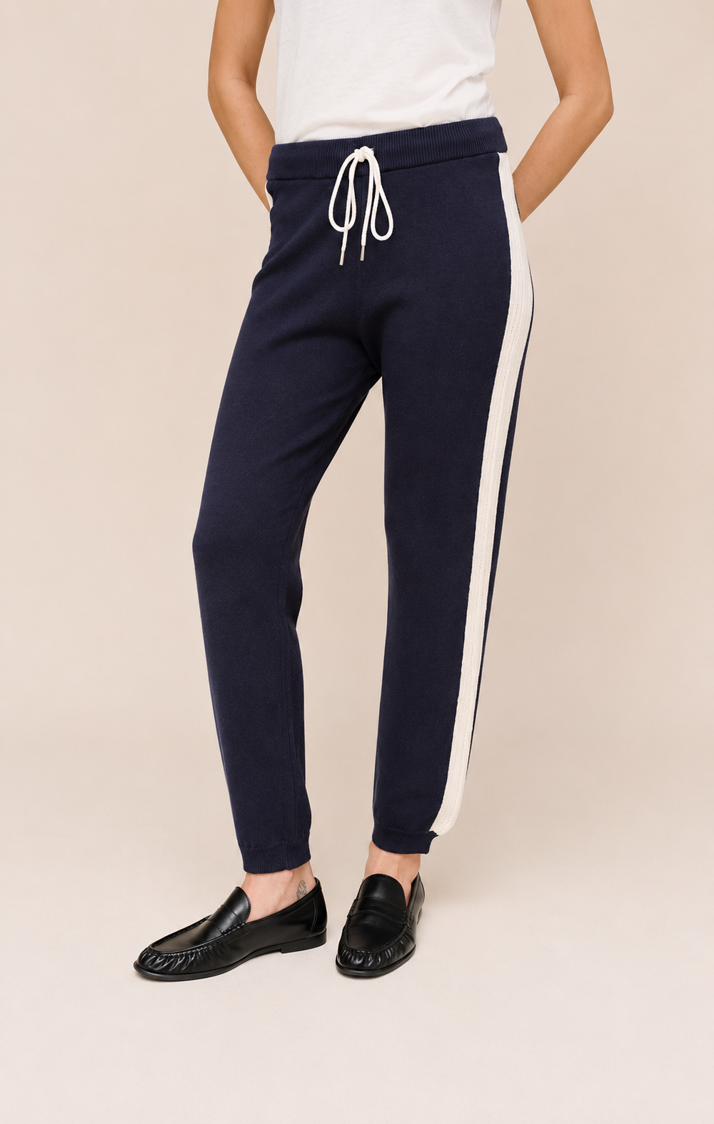 A person wearing navy jogger-style pants with a white side stripe and white drawstring, paired with a white top and black loafers, against a plain beige studio background.