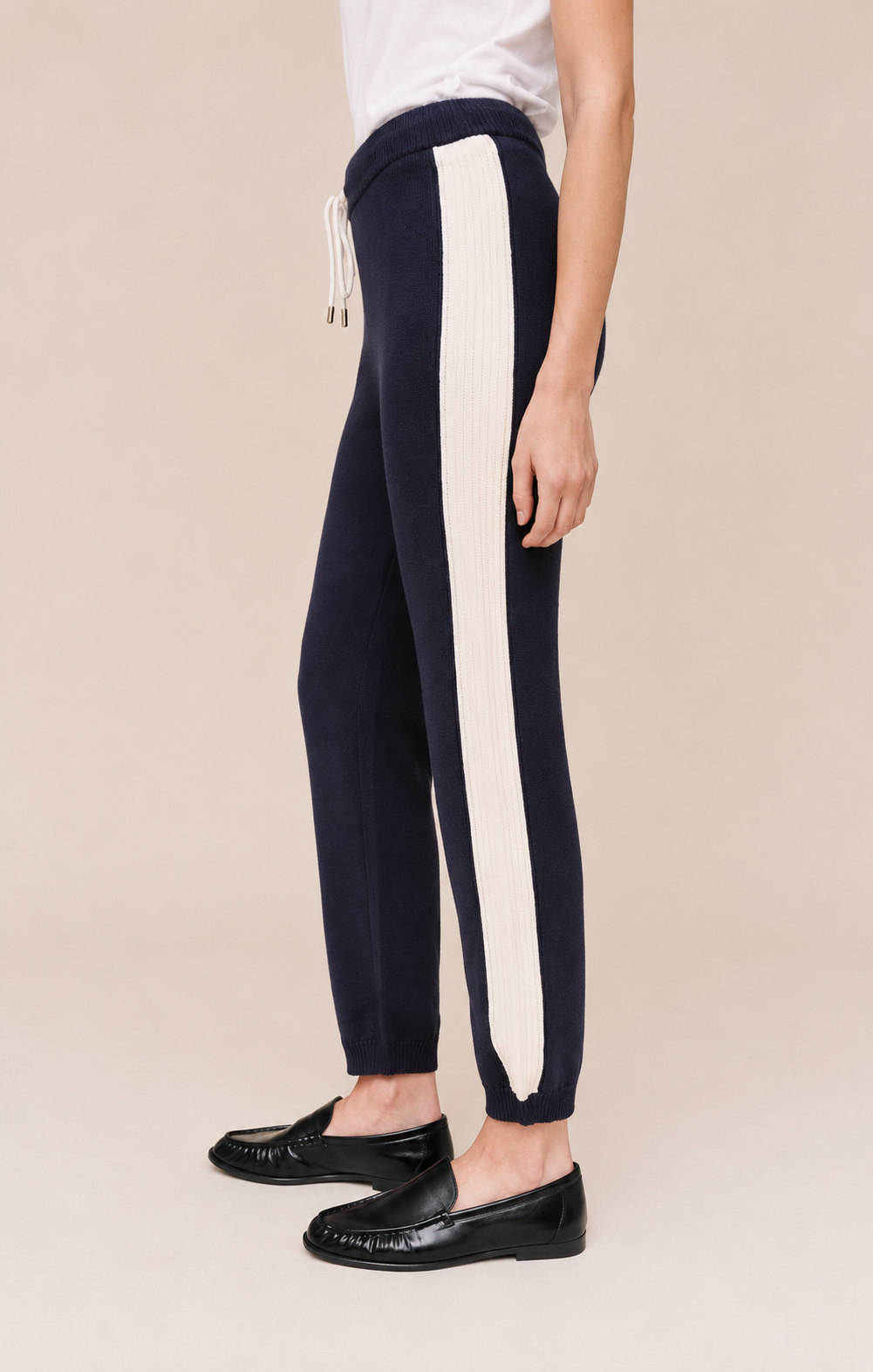 A person wearing navy jogger-style pants with a wide off-white vertical stripe down the side, paired with a white top and black loafers, against a plain beige studio background.